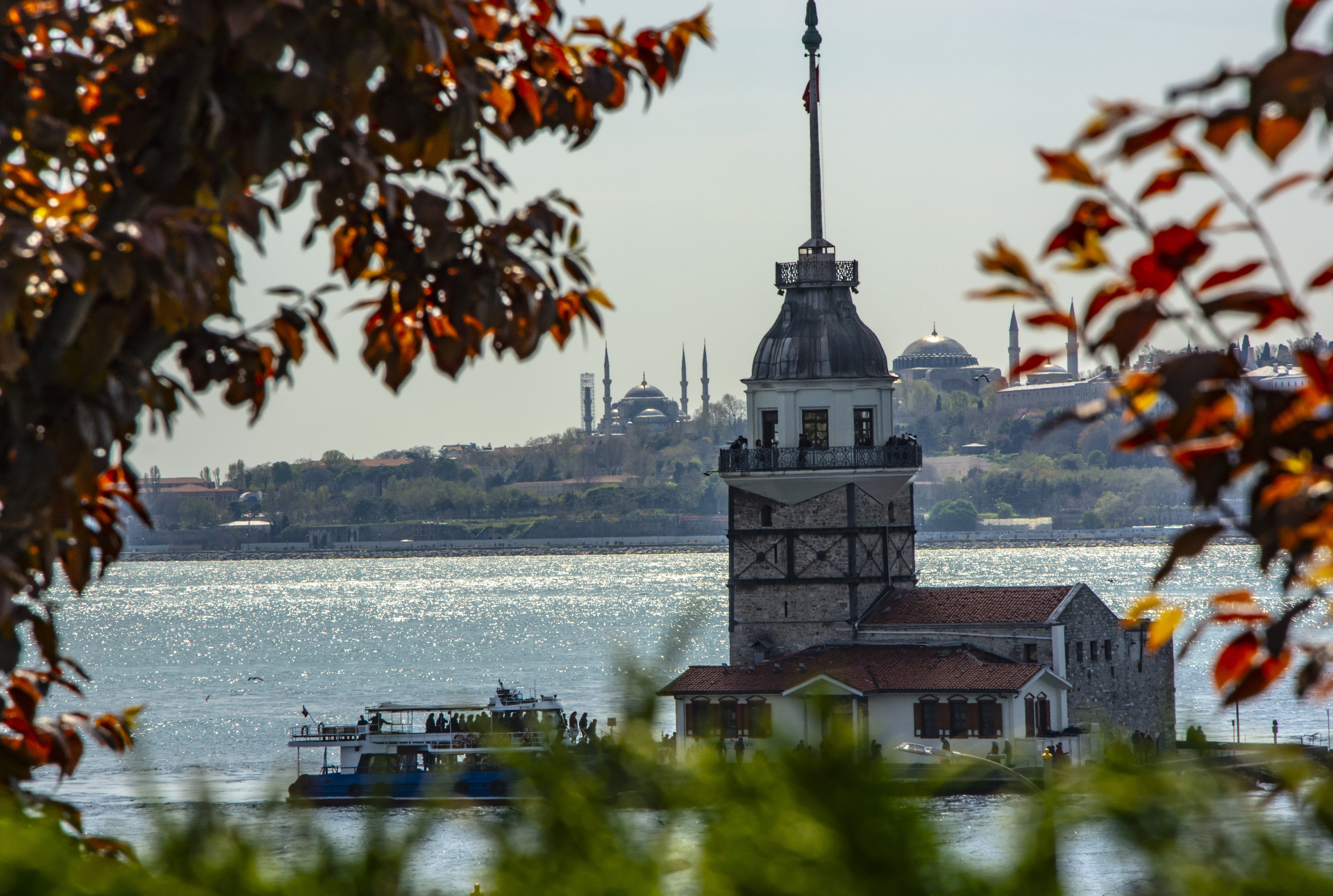 The Maiden's Tower in the Bosphorus with the historic peninsula skyline — Blue Mosque and Hagia Sophia — in the background, framed by autumn leaves